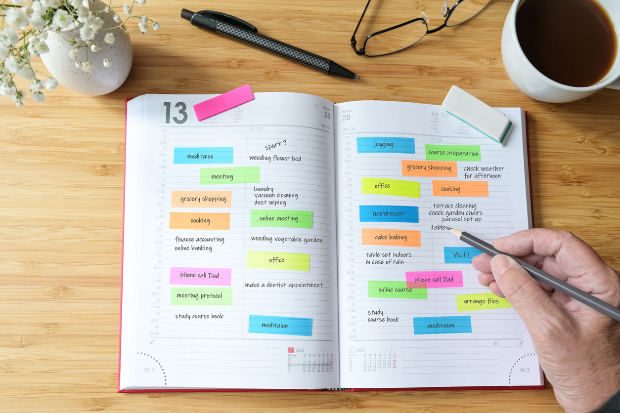 Hand of a woman writing appointments in a diary organizer or schedule calendar with a lot of colorful sticky notes for her time management on a wooden desk with coffee, flowers and glasses, selected focus